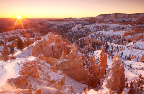 Sunrise over snow-covered rock formations and pine trees in a canyon landscape.
