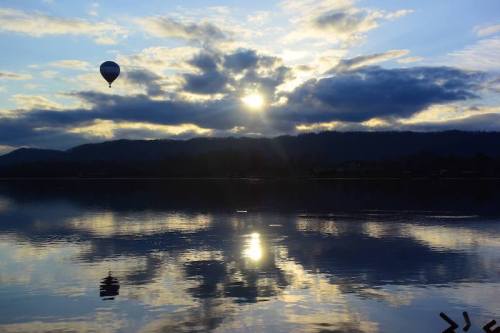 A hot air balloon floats above a lake at sunset, with clouds reflecting on the water's surface.