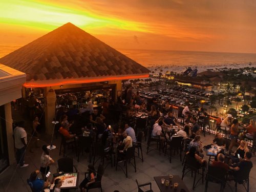 A vibrant sunset over the ocean, viewed from a rooftop bar filled with people enjoying drinks and food.