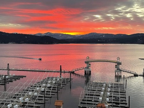 A vibrant sunset over a lake, with mountains in the background and a boat cruising past marina docks.
