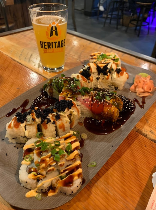 A platter of sushi rolls drizzled with sauces, garnished with green onions and pickled ginger, alongside a beer.