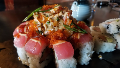 A close-up of sushi rolls topped with tuna, spicy sauce, green onions, and fish roe on a dark plate.