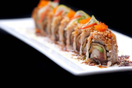 A close-up of a sushi roll topped with orange roe and garnished with vegetables, served on a white plate against a black background.