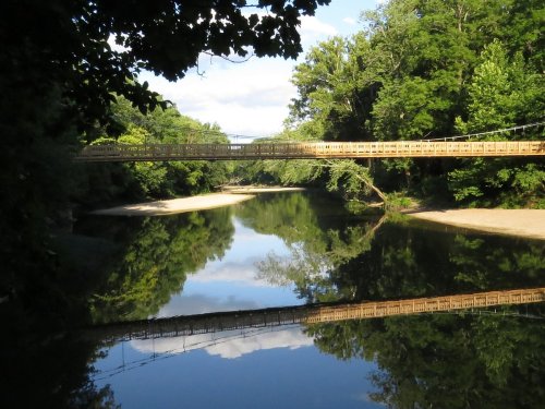A wooden bridge spans a calm river, surrounded by lush green trees and reflecting clouds in the water below.