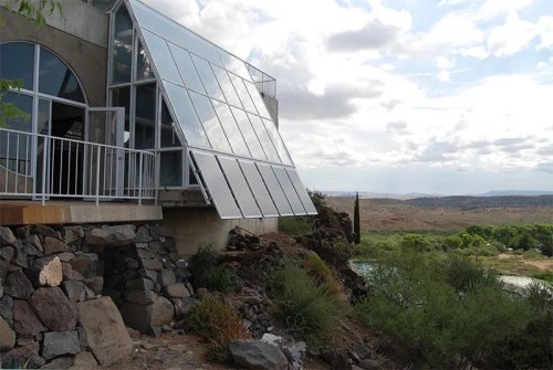 A modern house with large glass panels, set on rocky terrain near a river, surrounded by greenery and cloudy skies.