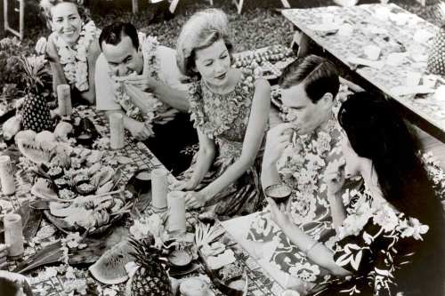 A group of five people enjoying a tropical feast, wearing leis and seated at a table adorned with fruits and decorations.