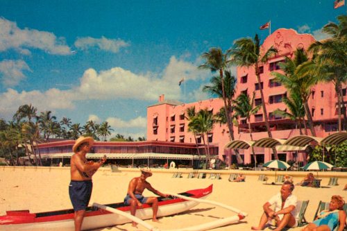 A vintage beach scene with a pink hotel, palm trees, and people enjoying the sun and water activities.