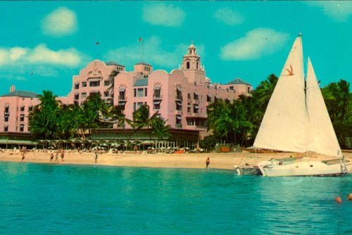 A vintage postcard of a pink hotel by the beach, with a sailboat in the foreground and palm trees lining the shore.