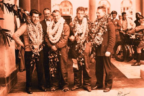 Five men in formal attire wearing flower leis, posing together outside a building with vintage vehicles in the background.