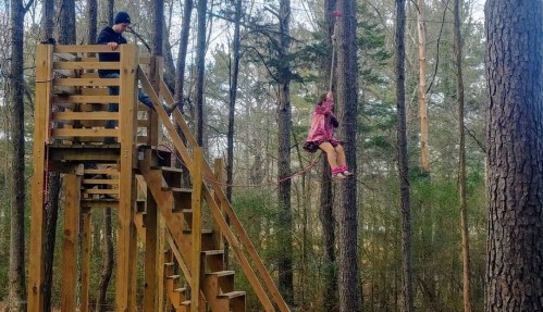 A child in a pink jacket sits on a zip line, while another person stands on a wooden platform in a forested area.