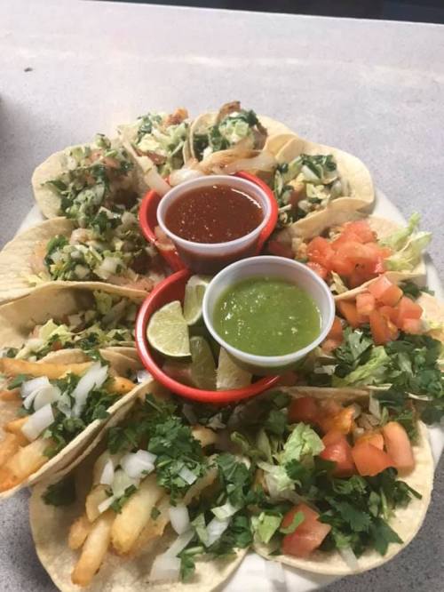 A platter of tacos with various toppings, served with red and green salsa, lime wedges, and fresh cilantro.