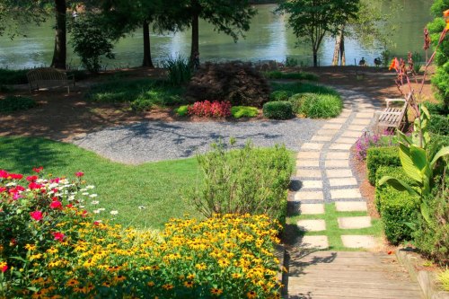 A serene garden with colorful flowers, a stone path, and a view of a river surrounded by trees.