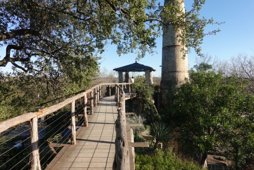 A wooden walkway leads to a gazebo near a tall stone structure, surrounded by trees and greenery under a clear blue sky.