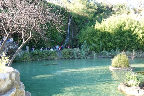 A serene natural scene featuring a waterfall, lush greenery, and people gathered near a tranquil blue-green pond.