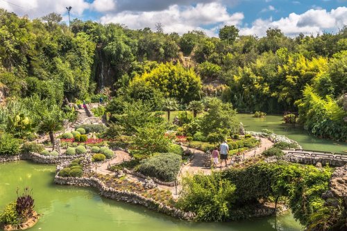 A scenic garden with lush greenery, pathways, and a tranquil pond under a blue sky with fluffy clouds.