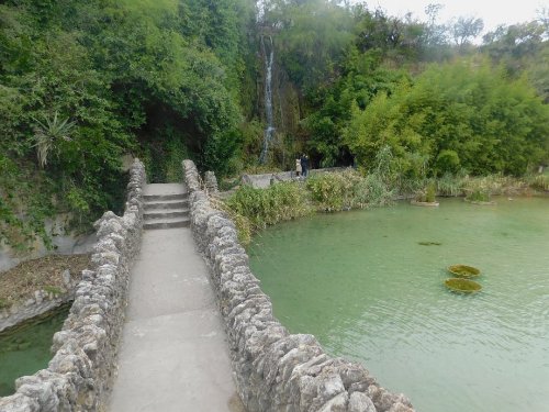 A stone bridge leads over a green pond, with a waterfall and lush greenery in the background.