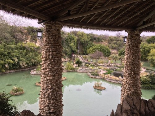 A serene garden with a pond, surrounded by lush greenery and stone pillars, under a thatched roof.