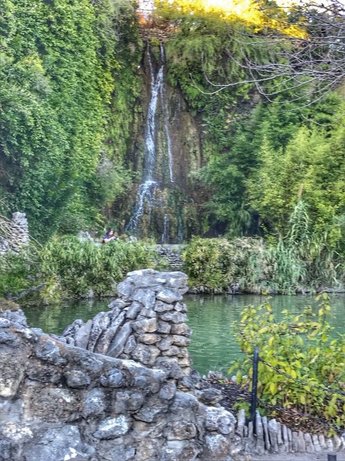 A serene waterfall cascades into a green pond, surrounded by lush vegetation and rocky formations.