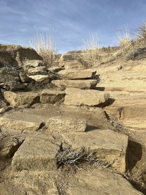 A rocky path leads upward, surrounded by dry grass and a clear blue sky.