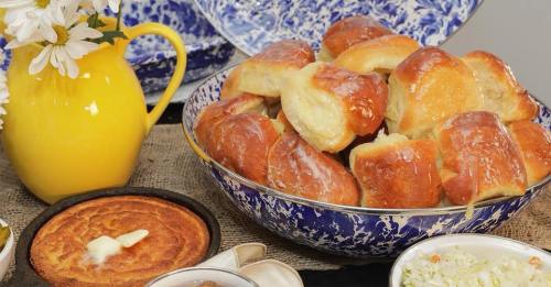 A bowl of golden, fluffy rolls with a yellow pitcher and side dishes of cornbread and coleslaw on a rustic table.