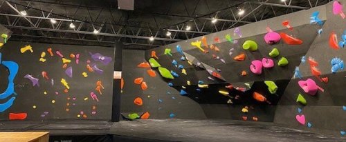 A vibrant indoor climbing gym with colorful holds on black walls and a padded floor.