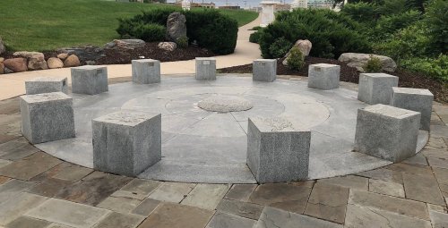 A circular stone monument with ten rectangular pillars arranged around a central circular design, set in a landscaped area.