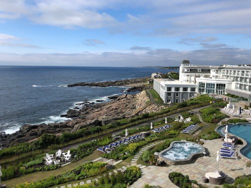 A coastal resort with a pool, gardens, and rocky shoreline under a cloudy sky. Waves gently crash against the rocks.