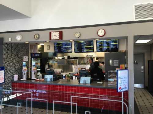 A fast-food counter with menus displayed, clocks on the wall, and staff serving customers in a casual setting.