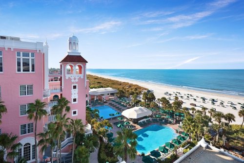 A pink hotel overlooks a pool area and beach, with palm trees and umbrellas under a clear blue sky.