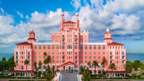 A large pink hotel with towers, surrounded by palm trees and a blue sky with clouds.