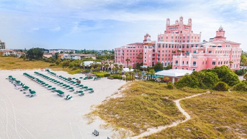 Aerial view of a pink hotel by the beach, with green lounge chairs on the sandy shore and palm trees in the background.