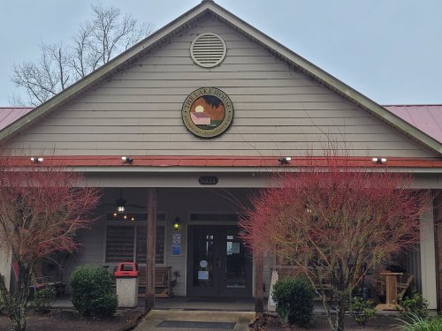 A rustic building with a circular sign, surrounded by shrubs and trees, on a cloudy day.