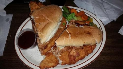 A plate with a sandwich made of fried fish, served with a side of mixed vegetables and a small cup of sauce.