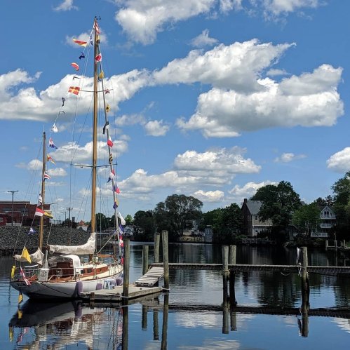 A sailboat adorned with colorful flags docked at a pier, surrounded by trees and a blue sky with fluffy clouds.