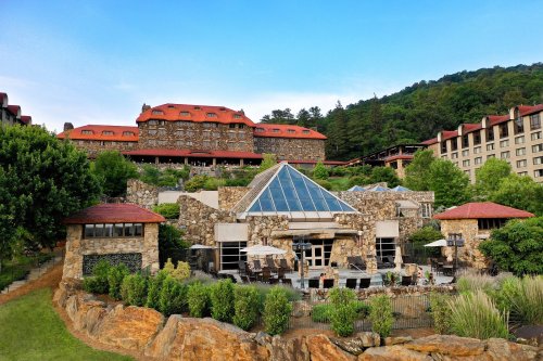 A scenic view of a stone resort with a glass atrium, surrounded by greenery and mountains under a clear blue sky.