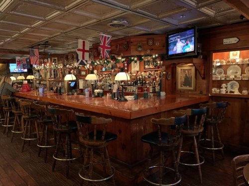 A cozy bar with wooden decor, flags, and a TV, featuring a long counter and high-backed stools.