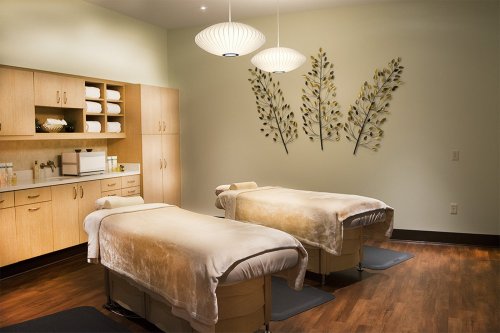 A serene spa room with two massage tables, soft lighting, and decorative wall art of leaves.
