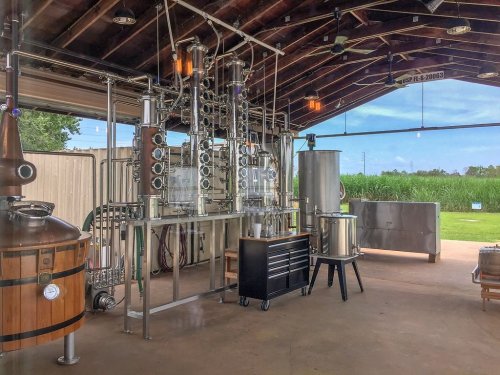 A distillery setup featuring copper stills, stainless steel tanks, and a spacious interior with a view of greenery outside.
