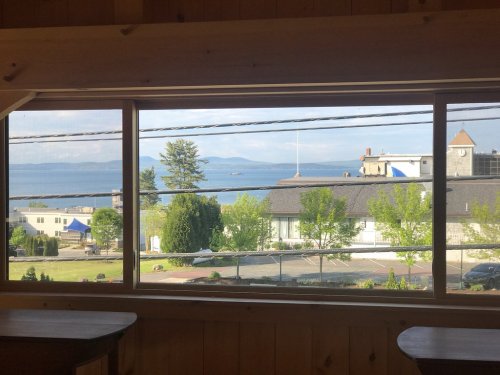 A scenic view of a calm lake and distant mountains framed by a wooden window, with greenery in the foreground.