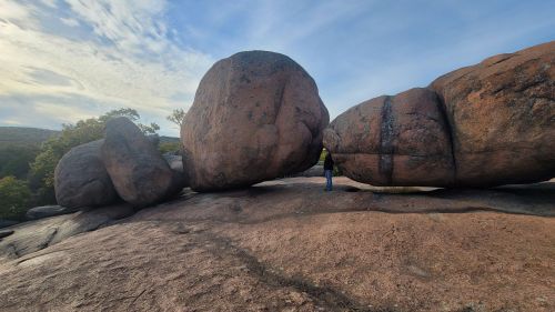 A person stands beside large boulders on a rocky surface under a clear sky, surrounded by trees in the background.