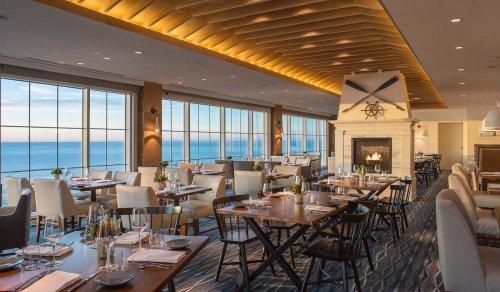 Elegant restaurant interior with large windows overlooking the ocean, featuring wooden tables and a cozy fireplace.