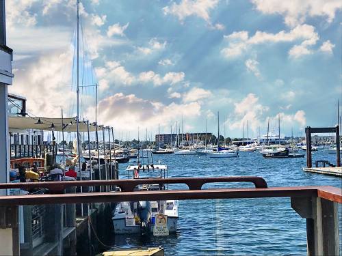 A scenic view of a marina with sailboats and yachts under a partly cloudy sky, seen from a waterfront restaurant.