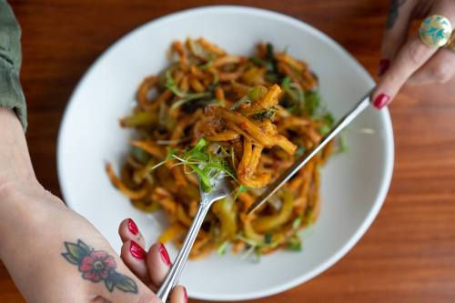 A close-up of a plate of pasta with greens, held by a hand with a tattoo, using a fork to serve.