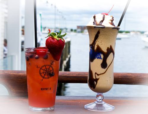 Two colorful cocktails sit on a wooden railing, with a scenic waterfront view in the background.