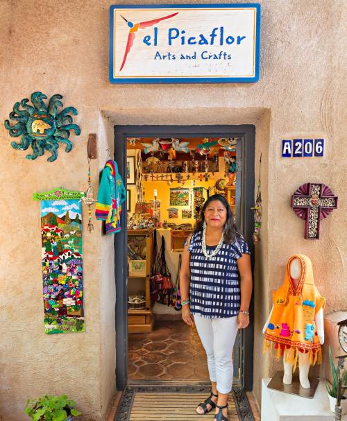 A woman stands at the entrance of "El Picaflor Arts and Crafts," surrounded by colorful decorations and crafts.