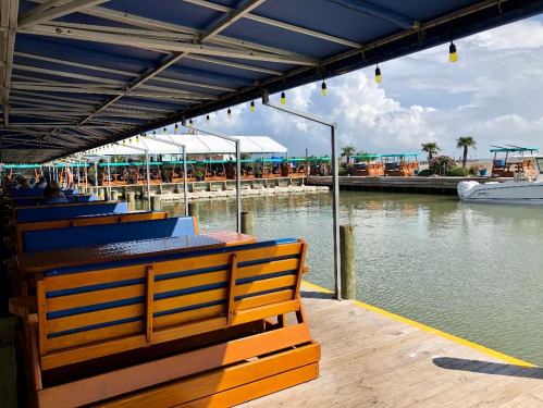 View of a waterfront dining area with wooden benches, boats, and a cloudy sky in the background.