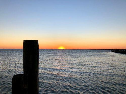 Sunset over calm water, with a wooden post in the foreground and a gradient sky transitioning from orange to blue.