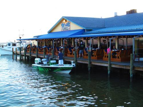 A waterfront restaurant with outdoor seating, a boat docked nearby, and diners enjoying the view.