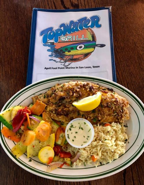A plate of grilled fish with lemon, rice, and sautéed vegetables, alongside a menu from Top Water Grill in Texas.