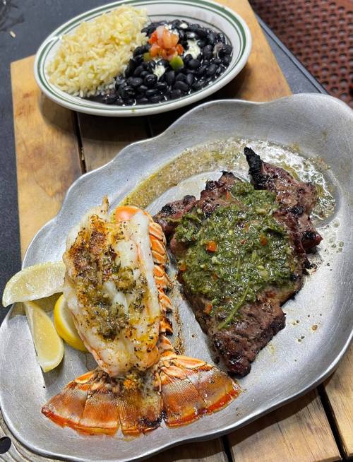 A silver platter with grilled lobster tail and steak topped with green sauce, served with rice and black beans in the background.